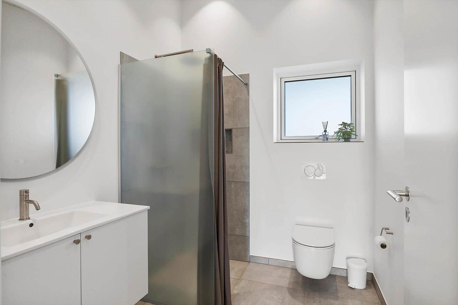 Badezimmer mit Dusche, Waschbecken und Toilette. Fenster mit Pflanze.