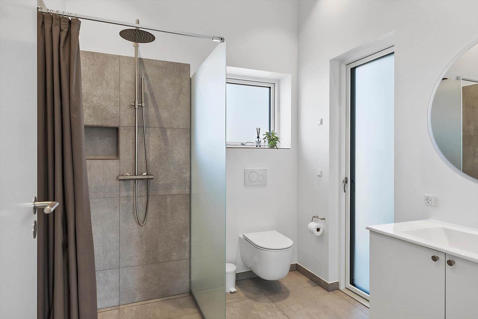 Badezimmer mit Dusche, WC und Waschtisch. Fenster und Spiegel sichtbar.