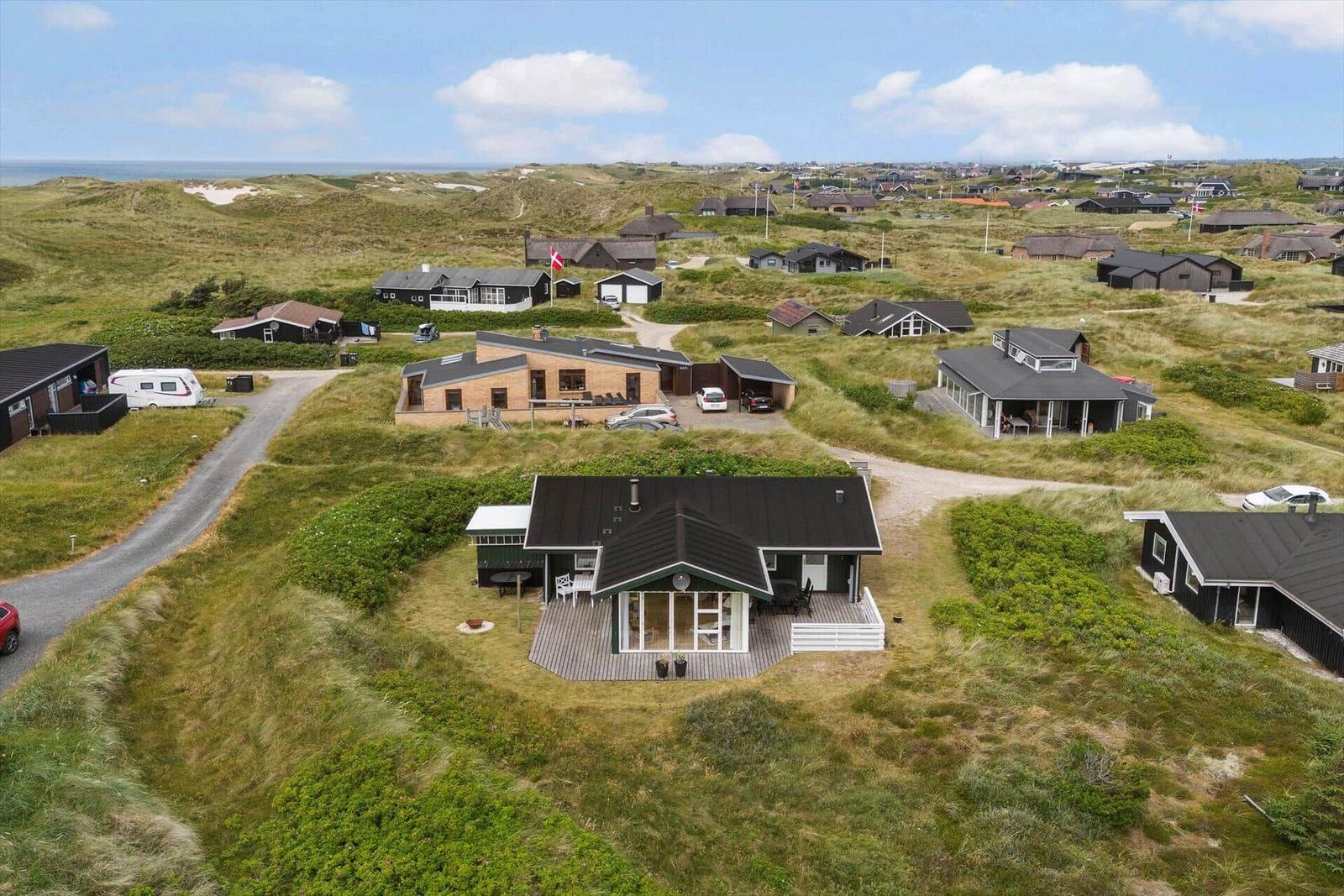 Haus mit Terrasse auf grüner Dünenlandschaft, Blick auf Meer und weitere Häuser.