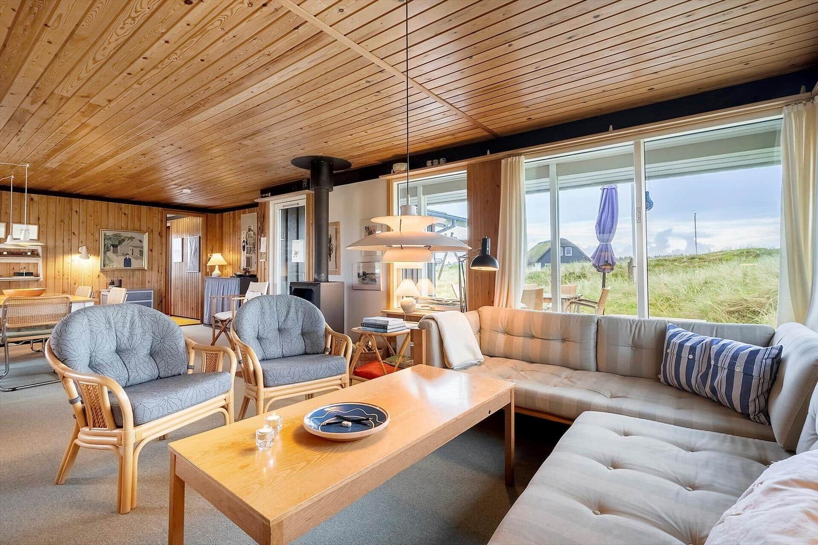 Wohnzimmer mit Sofa, Sesseln und Holzdecke. Große Fenster zeigen Landschaft.