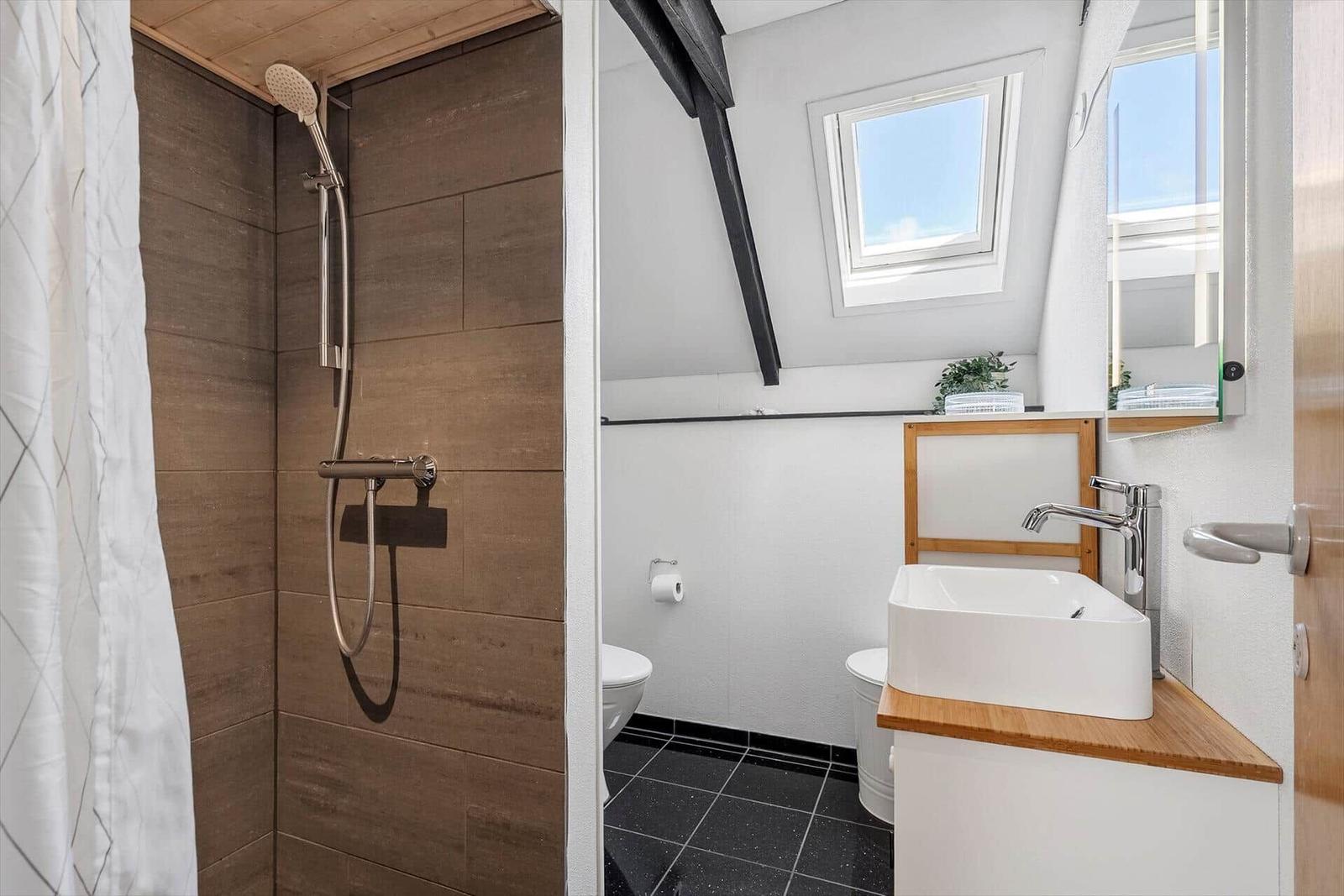 Shower with wood wall, sink and toilet under sloped roof with window.