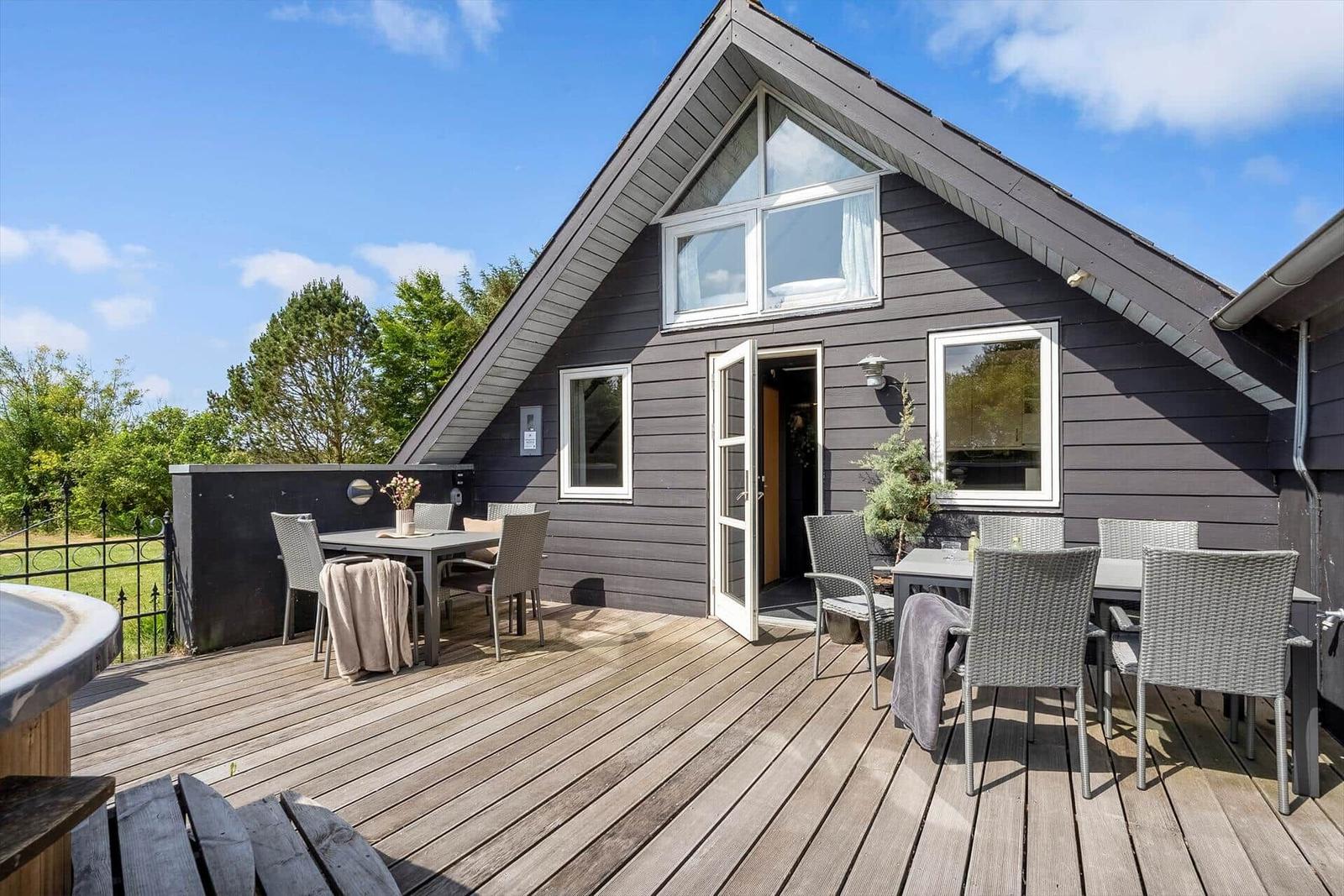 Wooden terrace with tables and chairs in front of a gray house with white windows.
