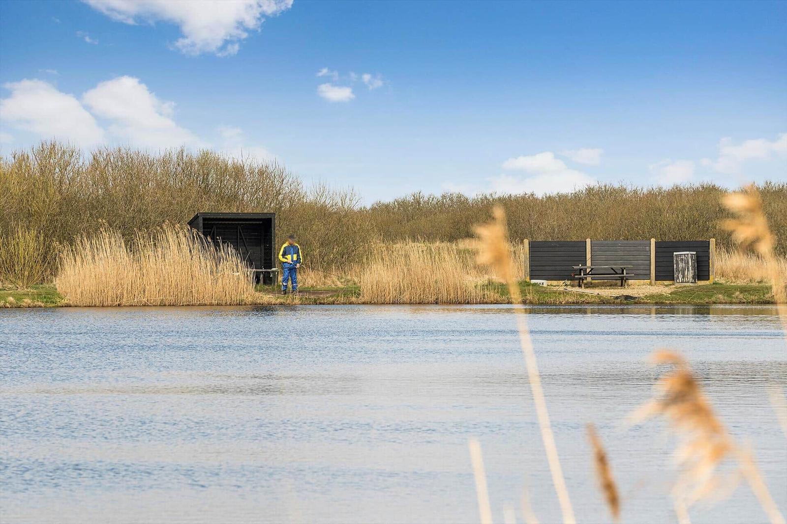 A fisherman stands by a lake. Trees and a wooden building are visible in the background.