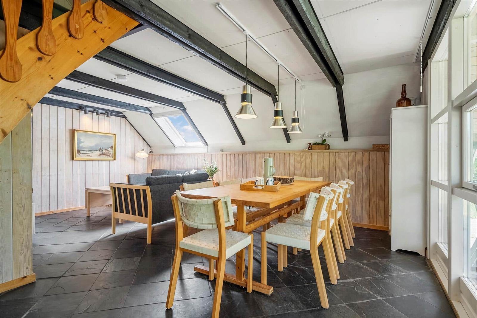 Dining room with wooden table, chairs, and sloped ceiling. Windows and sofa in the background.