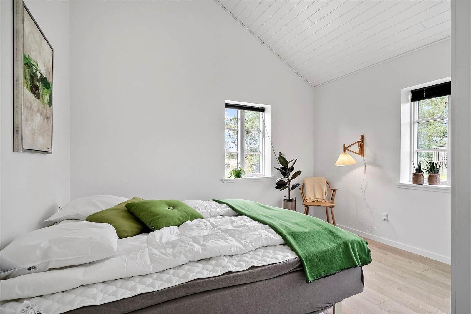 Schlafzimmer mit Bett, grünen Kissen und Decke, Fenster, Pflanzen und Wandlampe.