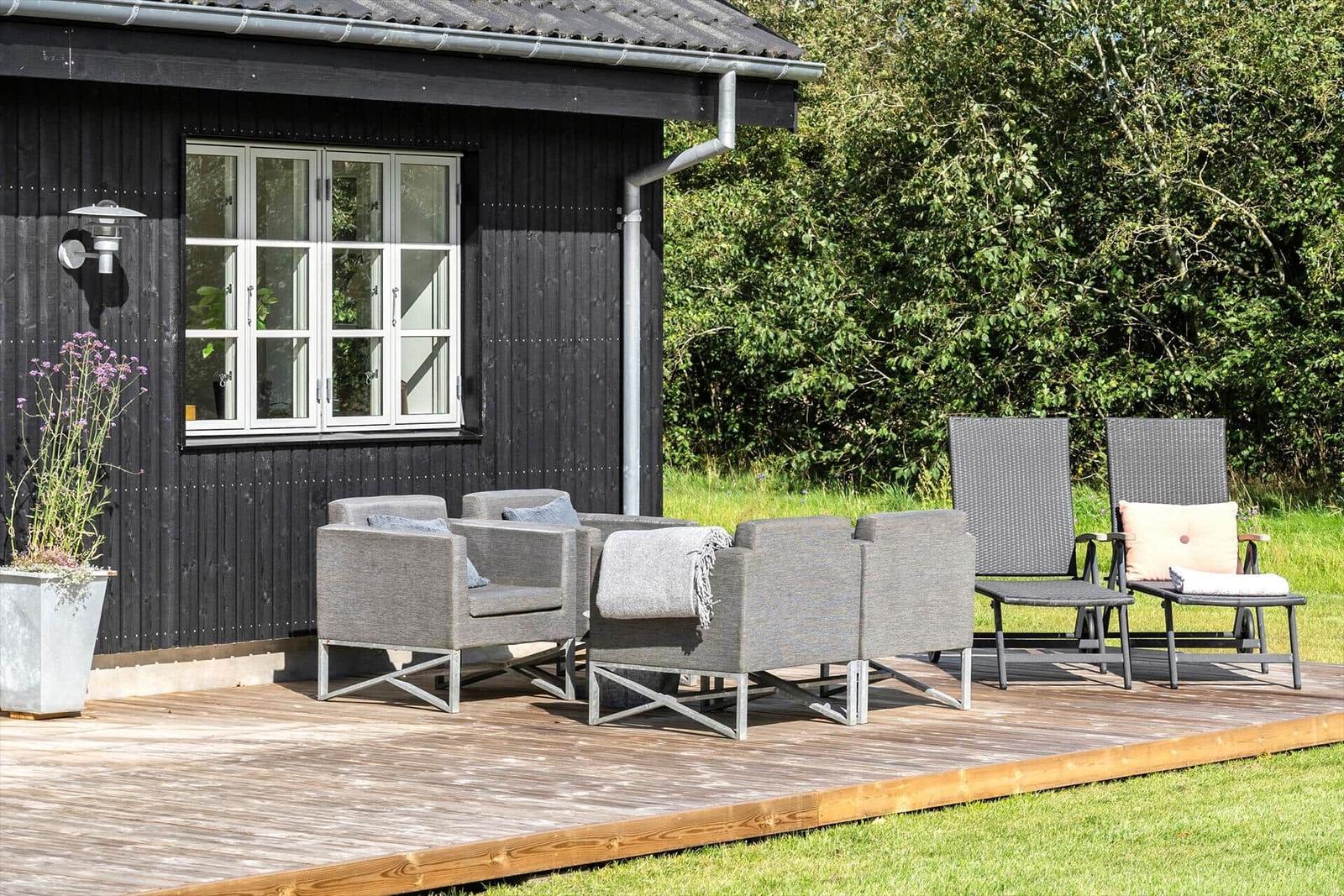 Terrasse mit Sitzgruppe und Liegestühlen vor schwarzem Holzhaus mit grünem Hintergrund.