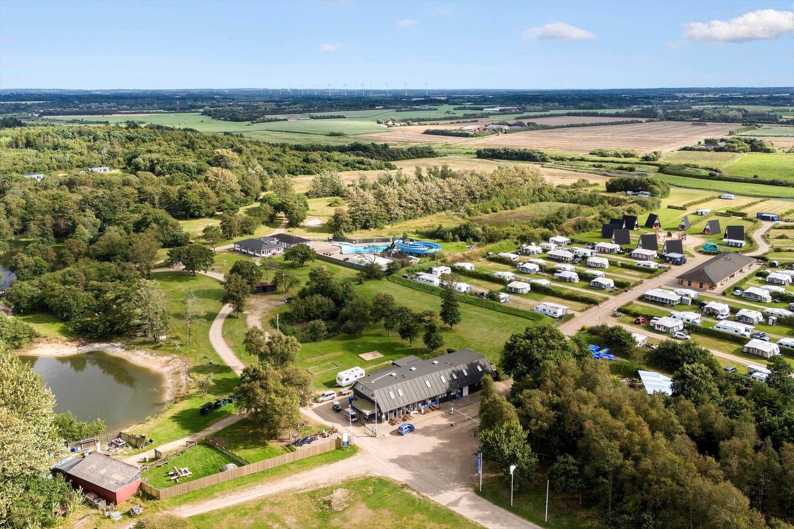 Grüner Campingplatz mit Pool, Wohnwagen und Wald. Blick auf Felder und Windräder.