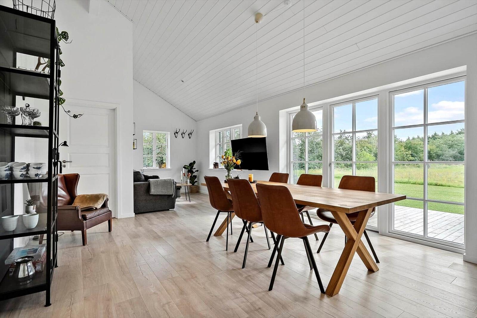 Esszimmer mit Holztisch und braunen Stühlen. Große Fenster zeigen Grünland. Wohnbereich im Hintergrund.