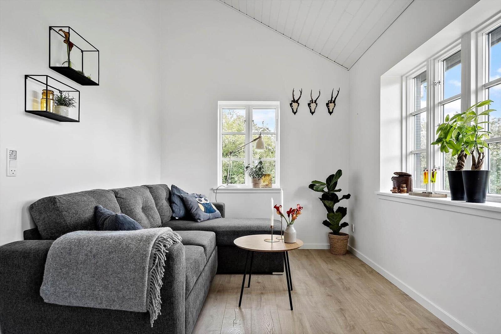 Wohnzimmer mit grauem Sofa, Holzboden und großen Fenstern. Wanddekoration und Pflanzen.