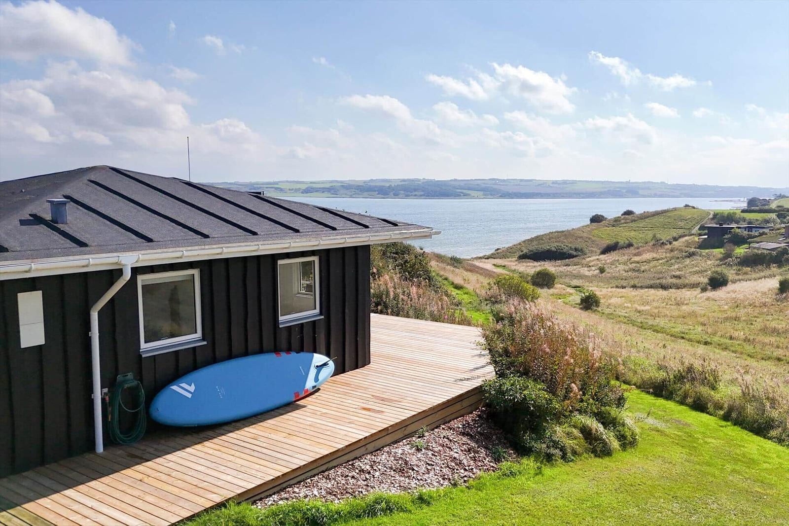 Haus mit Holzterrasse und Blick aufs Wasser. Blauer Surfbrett liegt bereit.