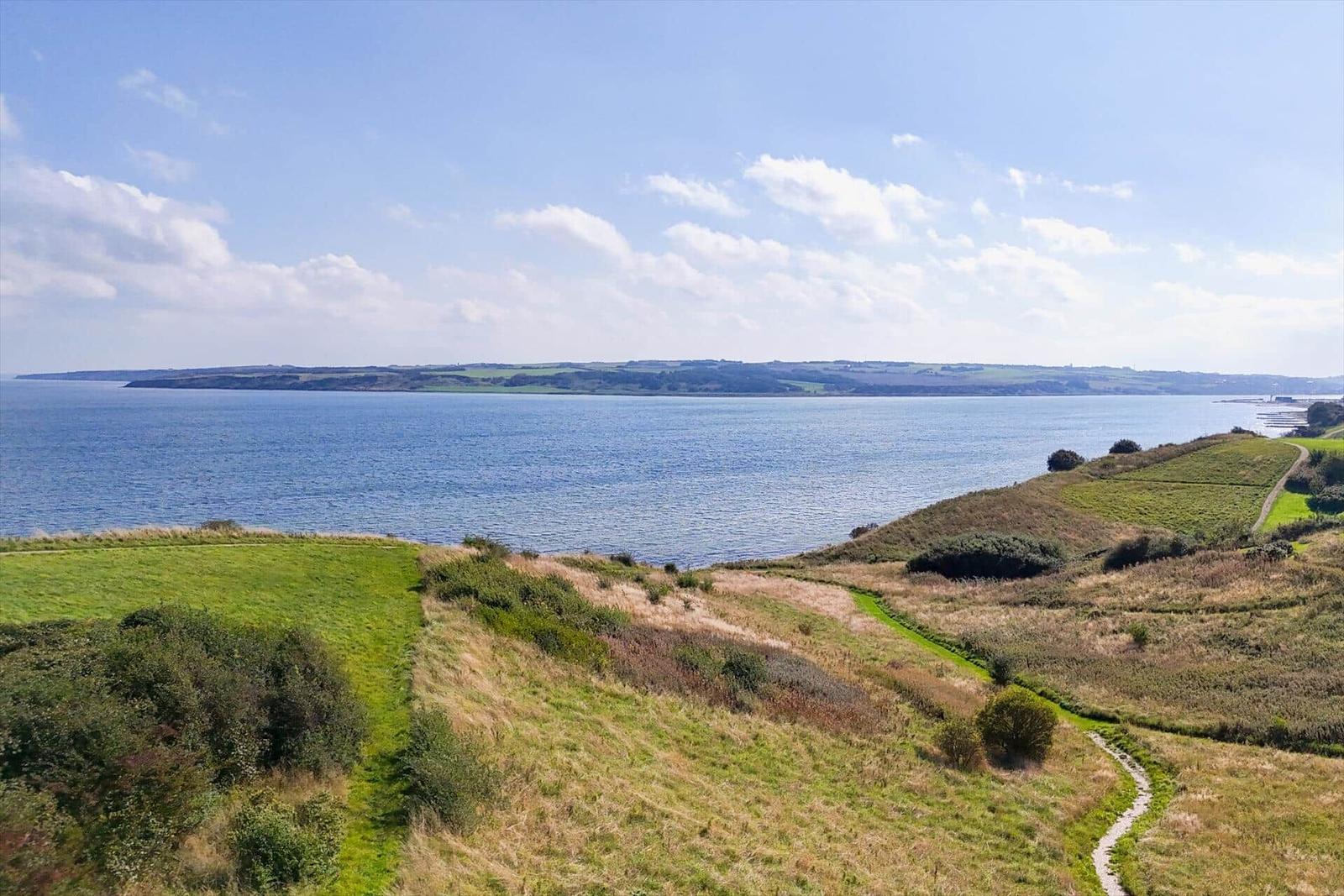 Weitläufiger Blick auf Küstenlandschaft mit Grünflächen und Wasser unter blauem Himmel.