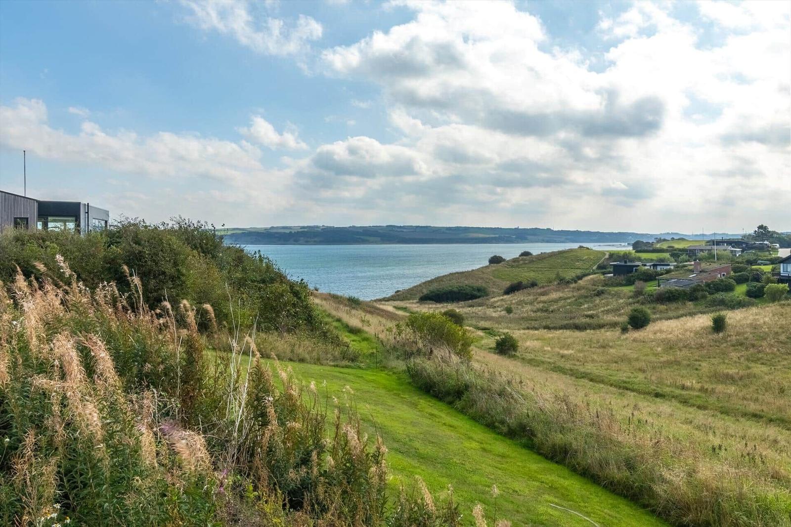 Modernes Haus mit Blick auf ruhige Küstenlandschaft und grüne Hügel unter blauem Himmel.