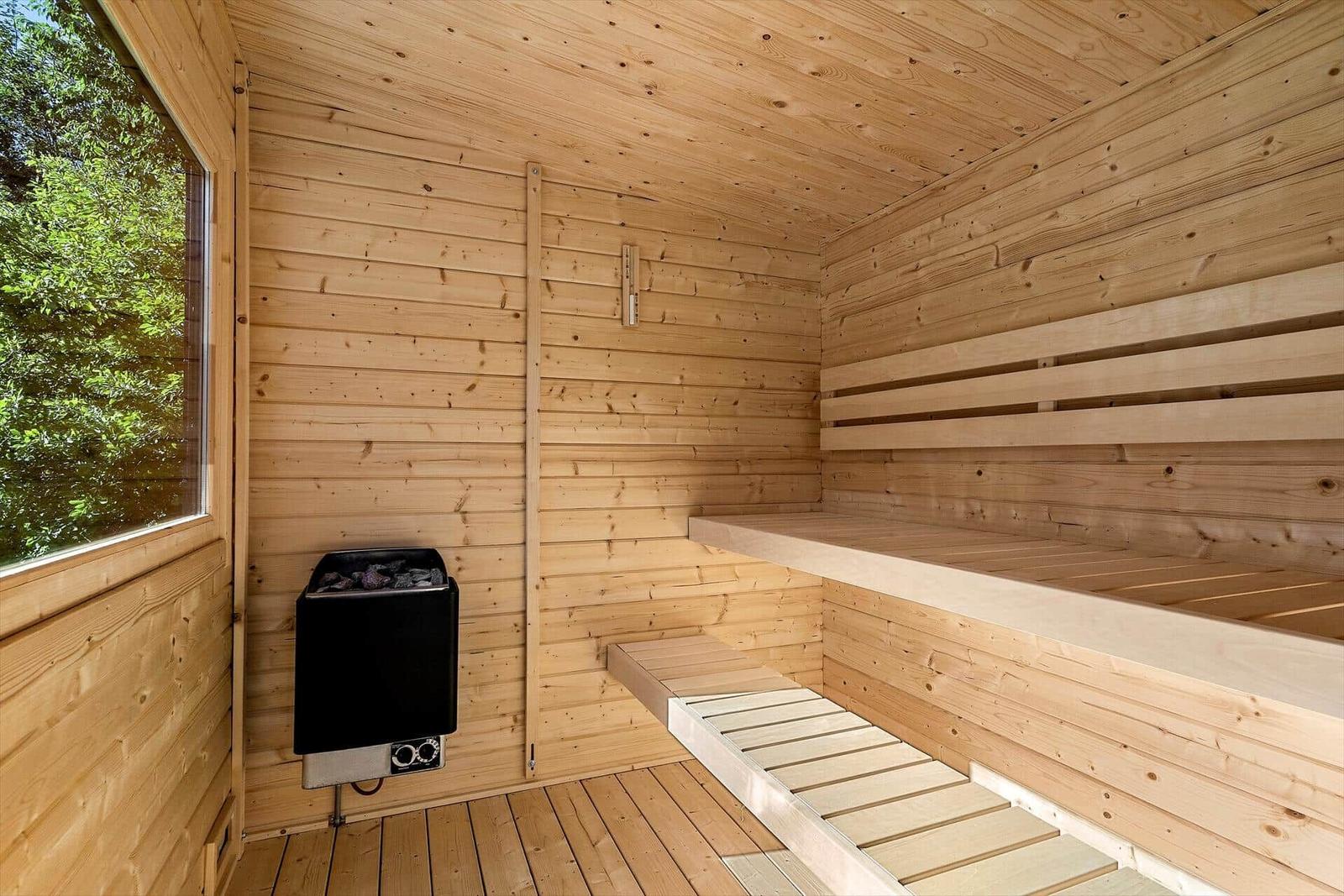 Sauna mit Holzwand, Bänken und Heizung. Fenster zeigt grüne Vegetation.