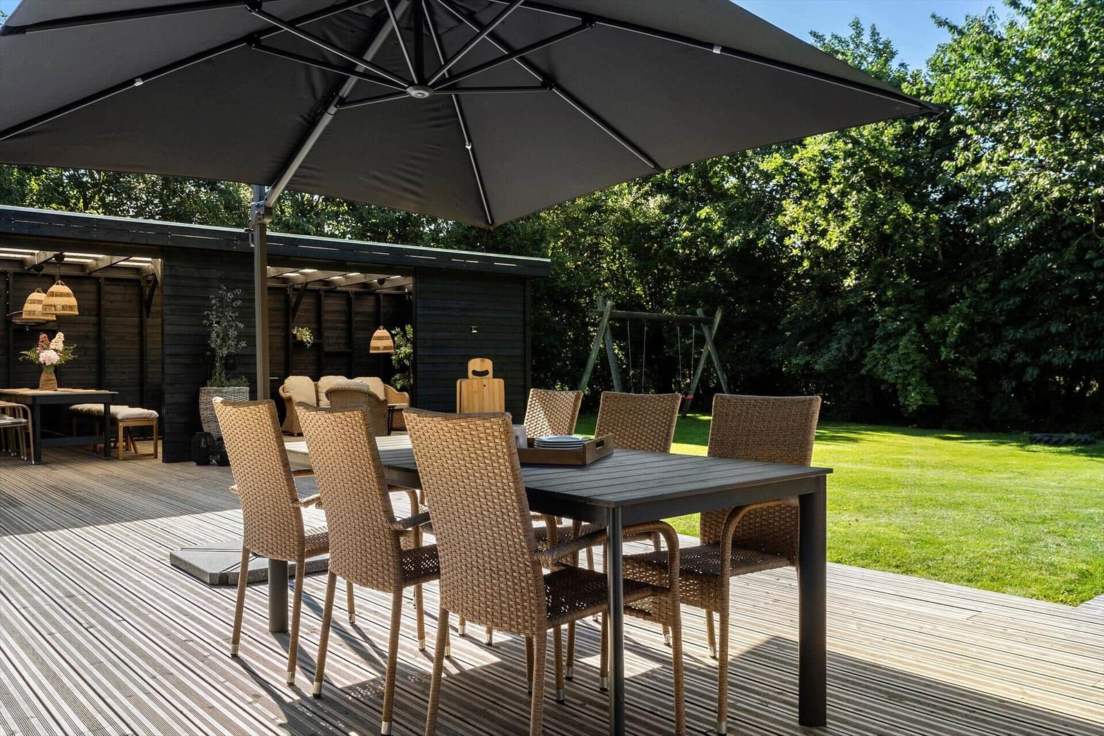 Terrasse mit Tisch, Stühlen und Sonnenschirm. Hintergrund: Garten und Holzhaus.