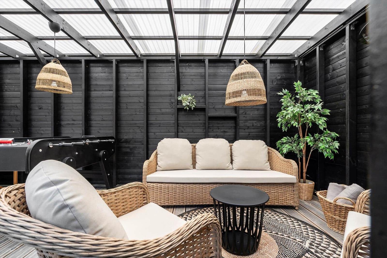 Terrasse mit Rattanmöbeln, Pflanzen und hängenden Lampen unter Glasdach.