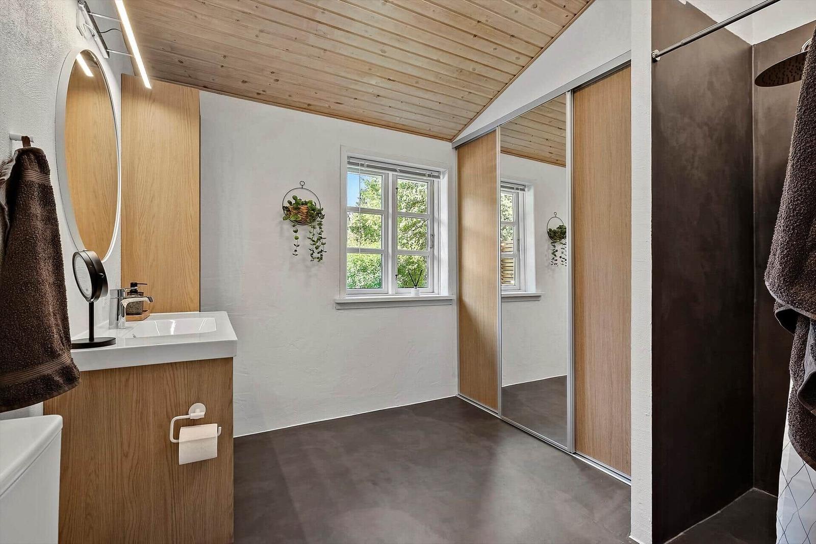 Badezimmer mit Holzdecke, Waschbecken, Dusche und Fenster mit Blick auf Grün.