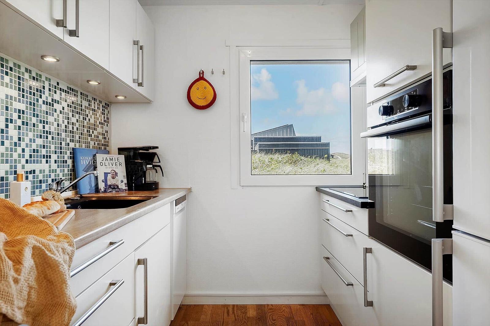 Küche mit weißen Schränken, Mosaikwand und Fensterblick auf Dünenszene.