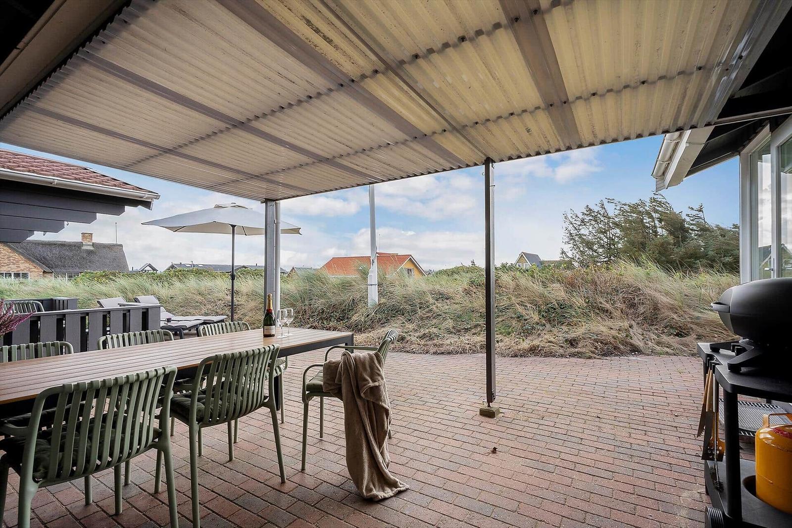 Terrasse mit Tisch, Stühlen und Grill unter Dach. Blick auf Dünen und Häuser.