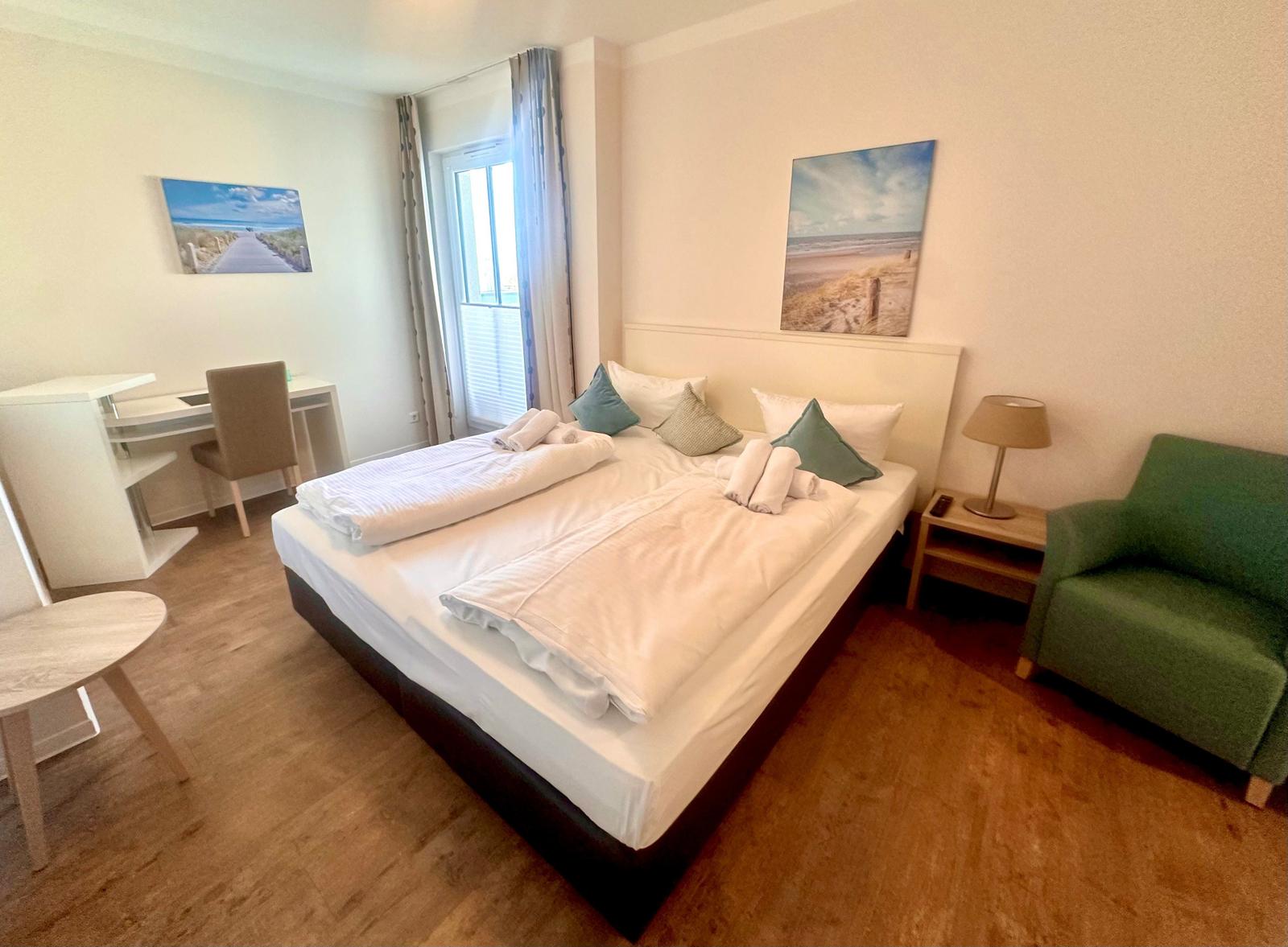 Schlafzimmer mit Bett, Schreibtisch und Sessel. An der Wand hängen Bilder.