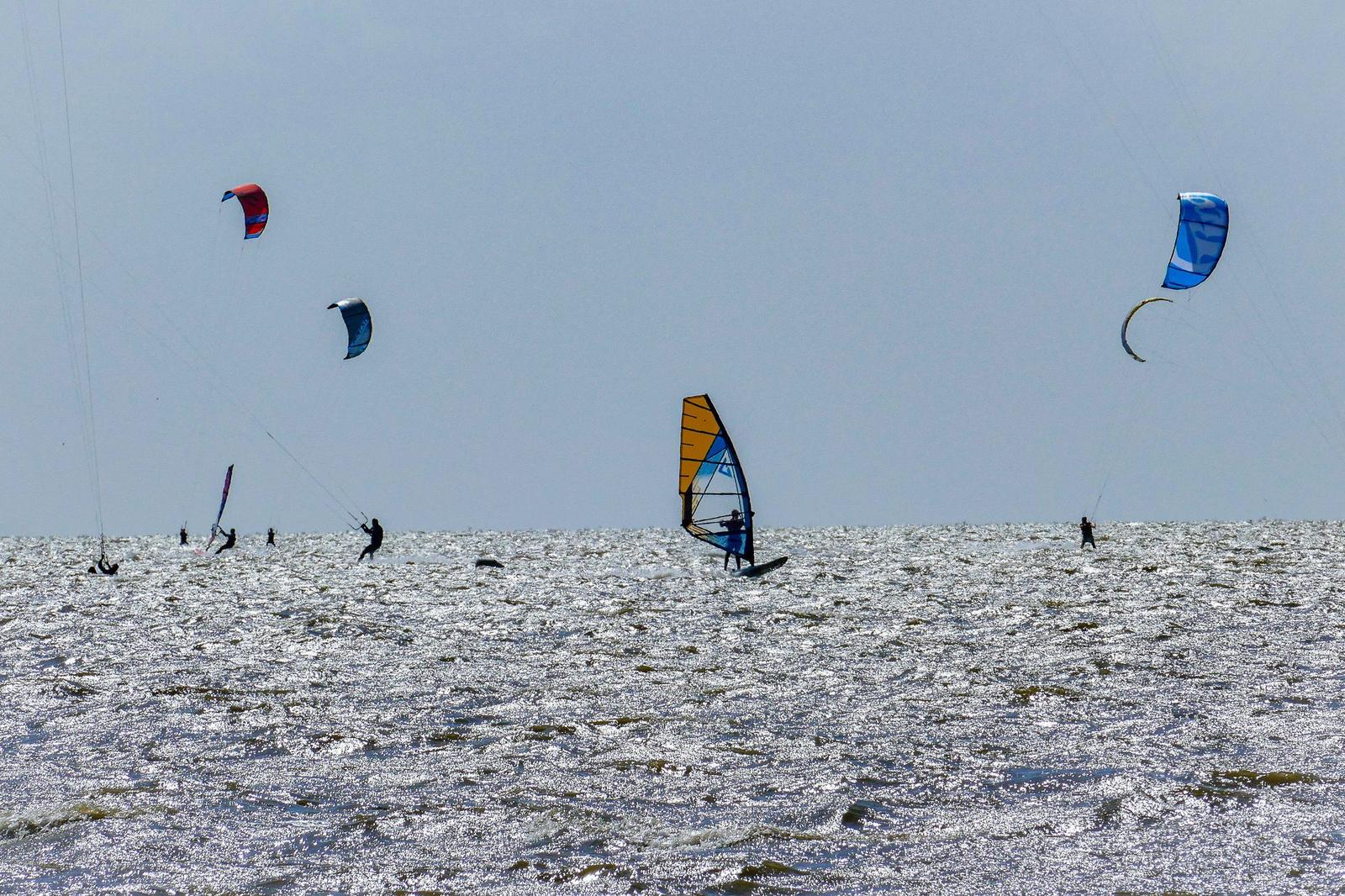 Multiple kitesurfers and windsurfers on the water with colorful sails.