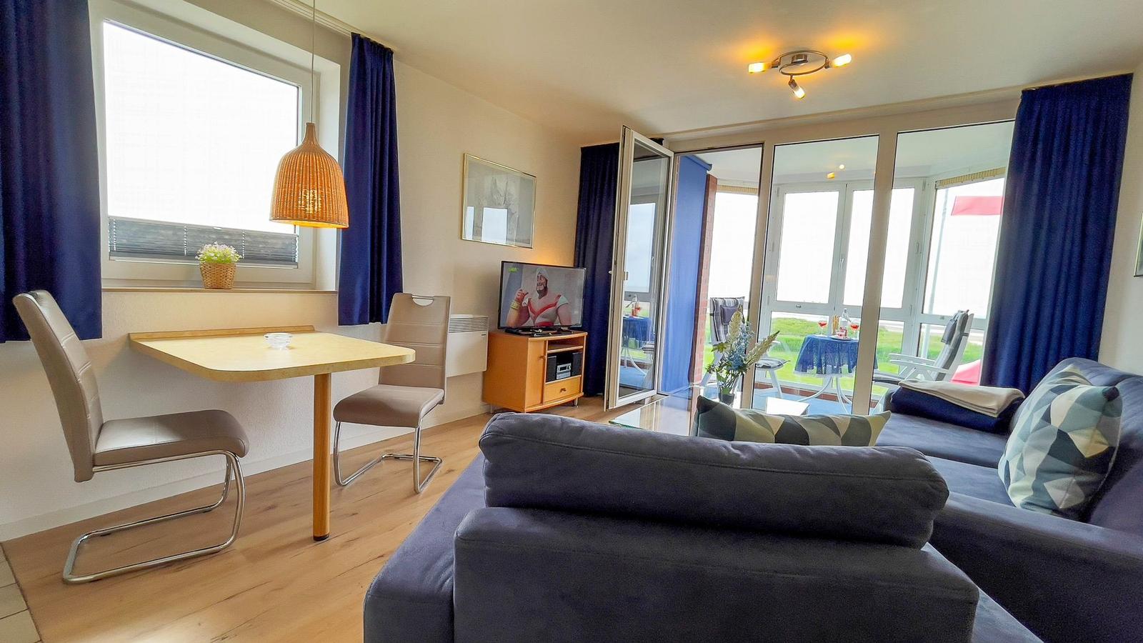 Living area with sofa, dining table, and TV. Entrance to terrace with garden view.