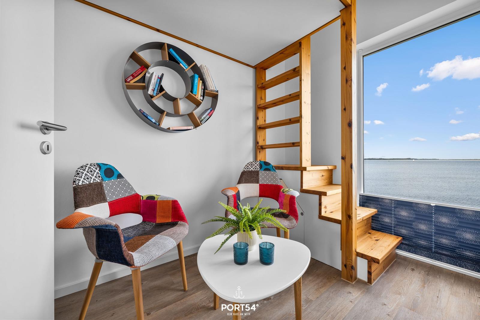 Zimmer mit Blick auf Wasser, zwei Stühle, Tisch und Bücherregal an der Wand.