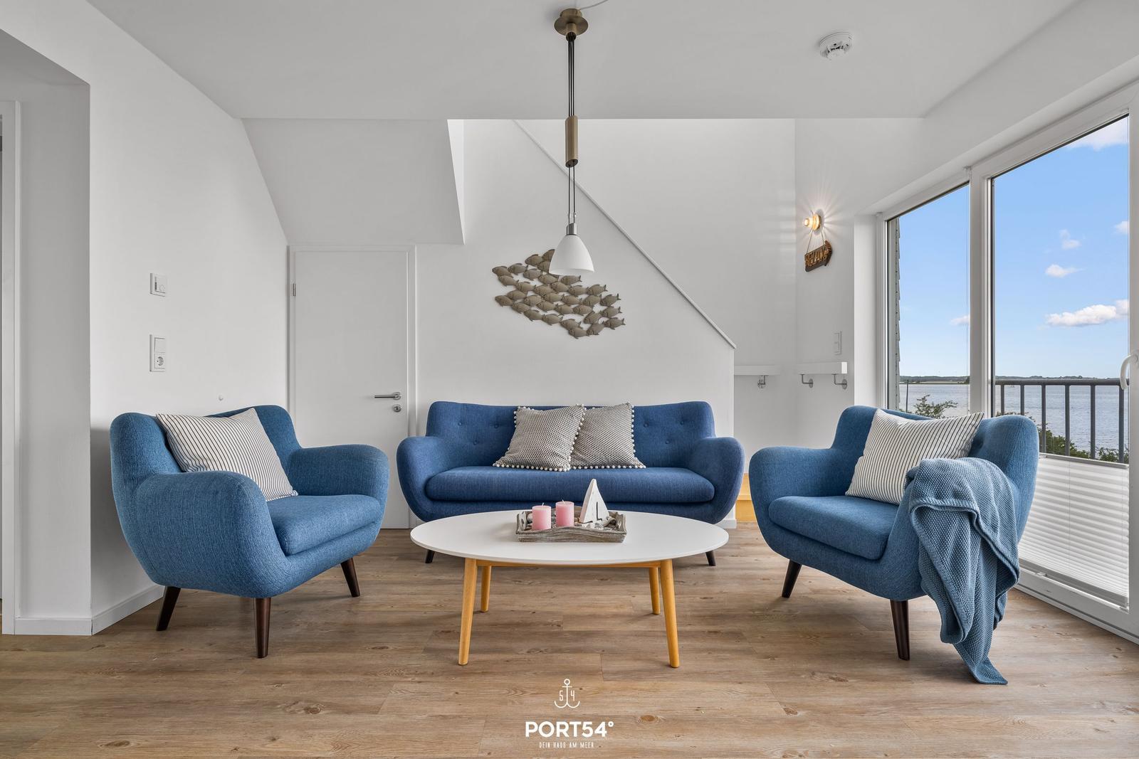 Wohnzimmer mit blauer Couch und zwei Sesseln. Blick auf Wasser durch große Fenster.