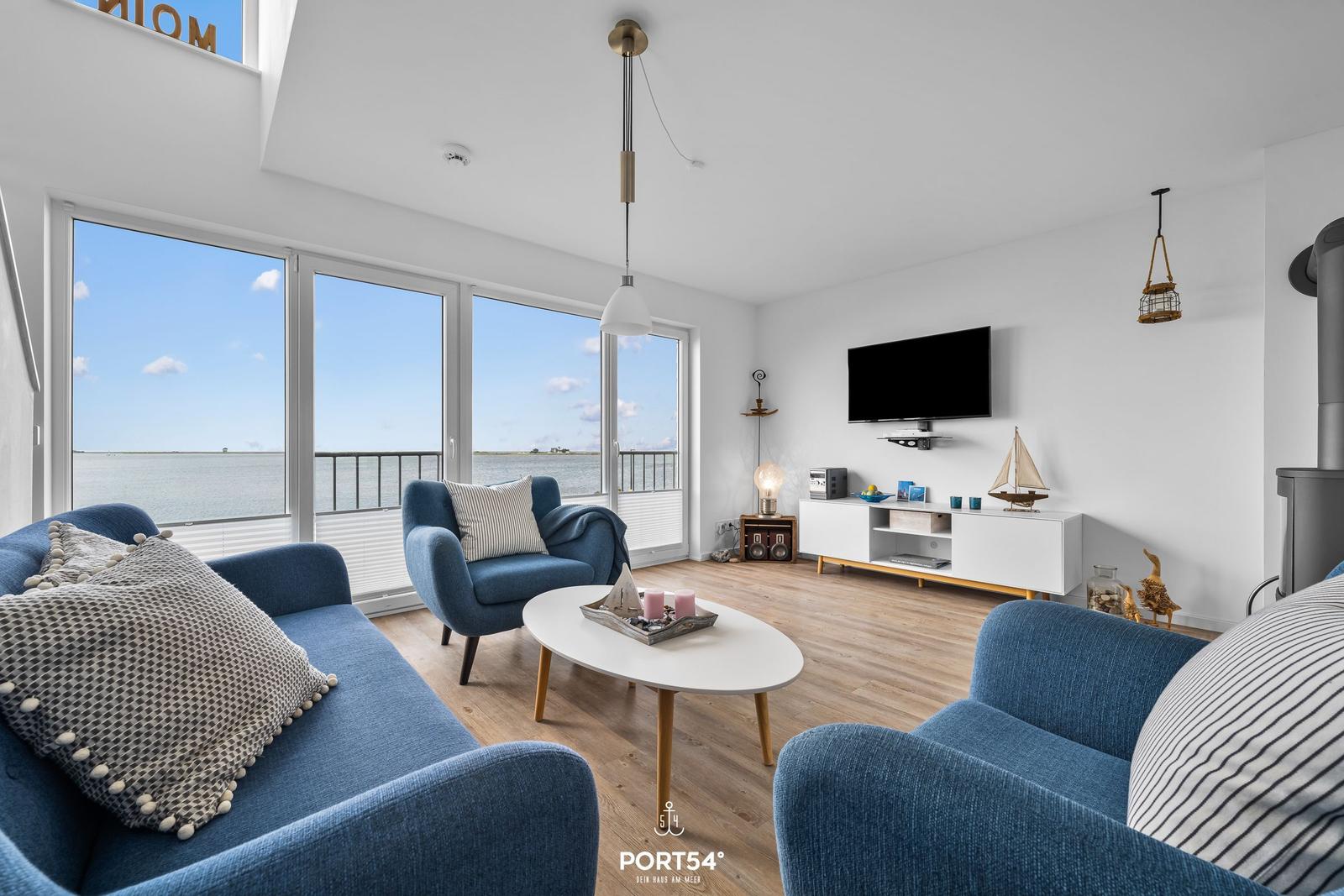 Wohnzimmer mit blauen Sesseln, TV und Meerblick durch große Fenster.