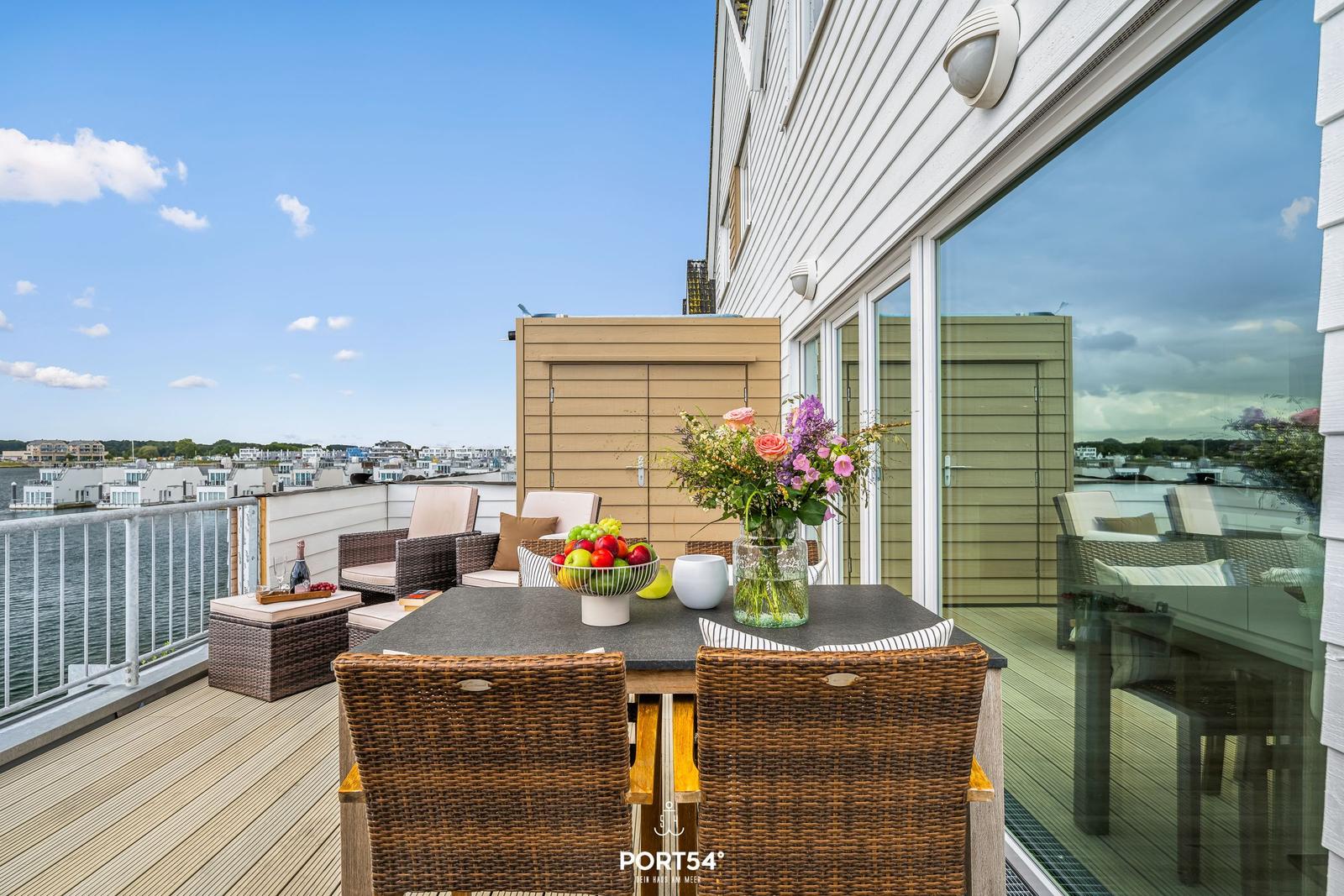 Terrasse mit Tisch, Stühlen und Blick auf Wasser und Gebäude.