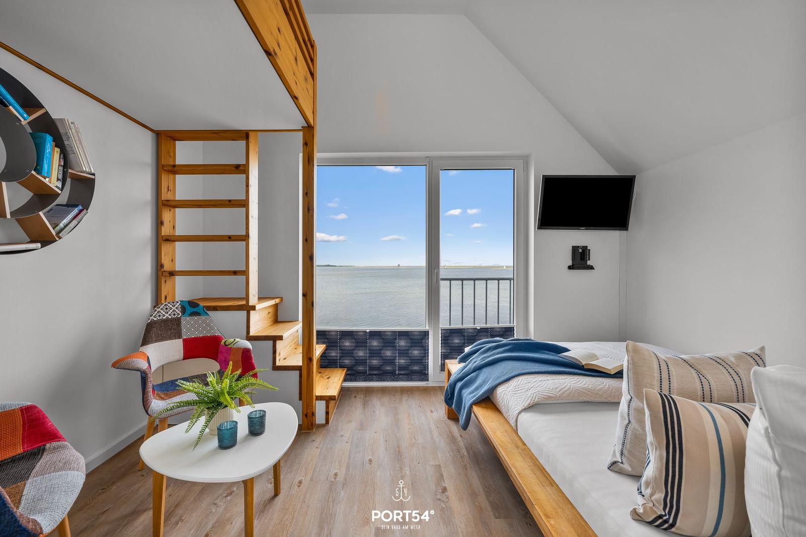 Schlafzimmer mit Blick aufs Wasser, Bett, TV und Holztreppe.