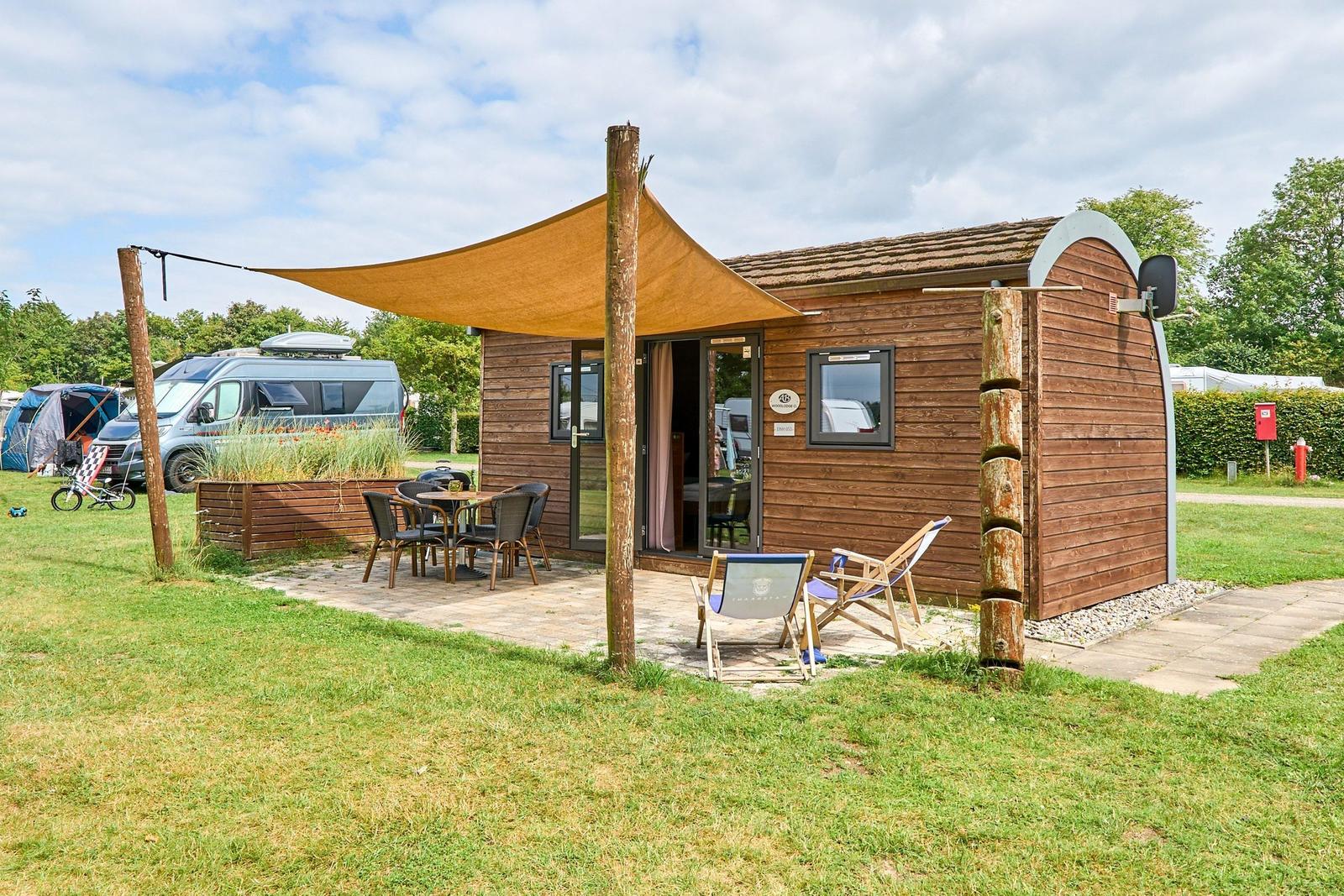 Holzhütte mit Terrasse, Sonnensegel und Gartenmöbeln. Hintergrund mit Wohnmobil und Zelt.
