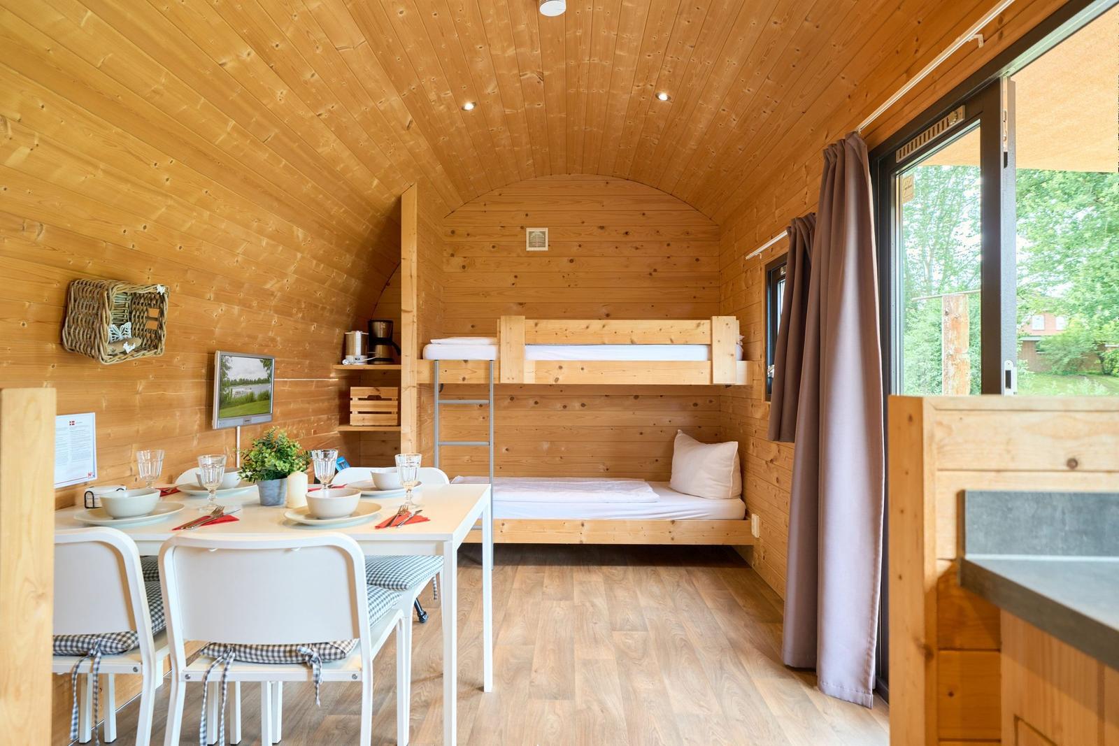 Holzhaus mit Schlafcouch, Esstisch und TV. Fenster mit Blick auf Grün.