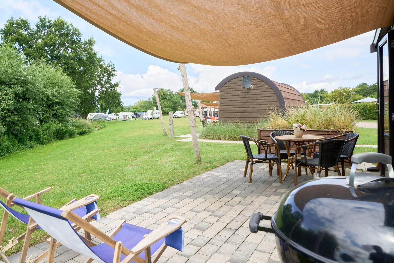 Terrasse mit Sitzgruppe, Grill und Sonnensegel. Grünfläche und Hütte im Hintergrund.