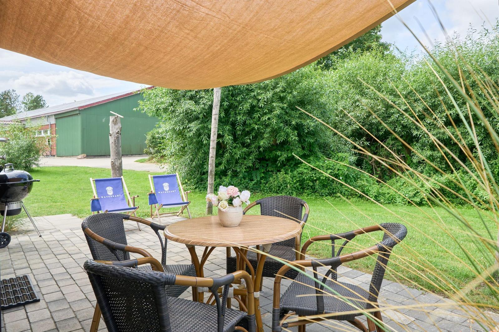 Terrasse mit Tisch, Stühlen, Sonnensegel und Grill. Hintergrund: grüner Garten und Gebäude.