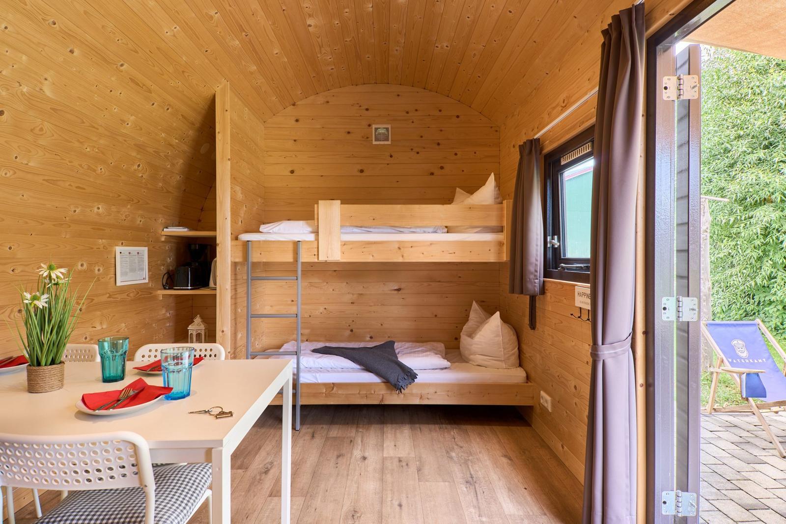 Holzvertäfelte Unterkunft mit Doppelstockbett und Tisch mit Stühlen. Durchgang zur Terrasse.