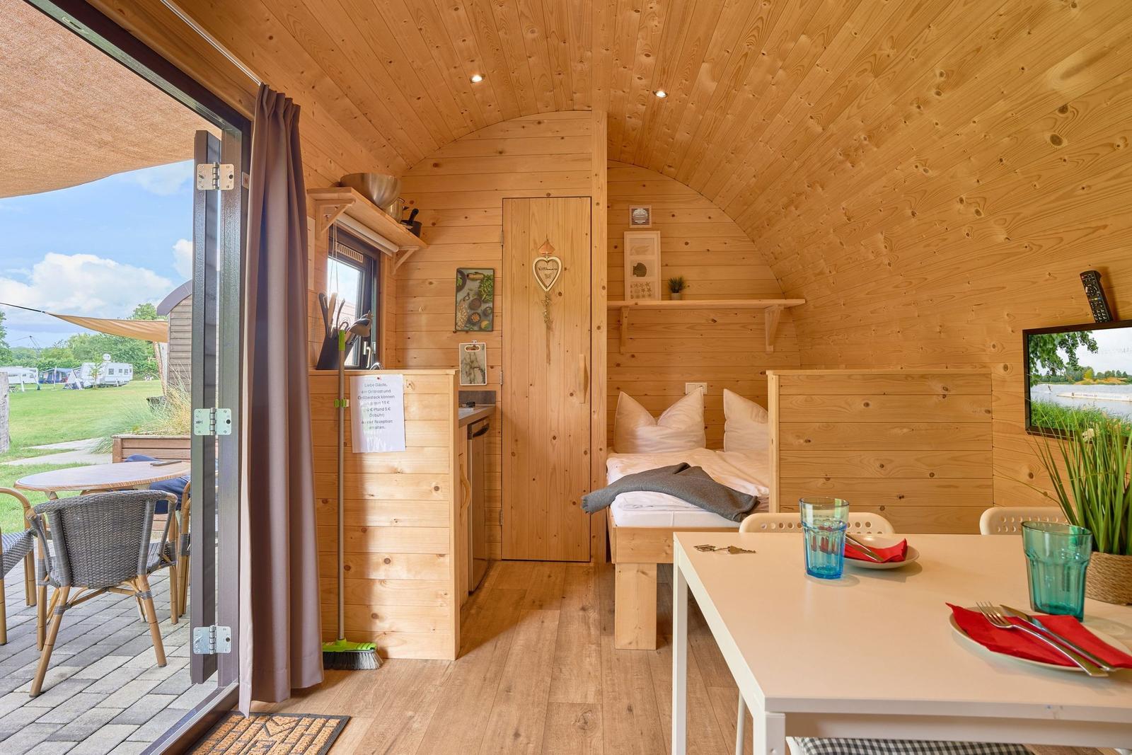 Holzverkleidete Unterkunft mit Schlafplatz, Esstisch und Terrasse mit Blick auf Campingplatz.