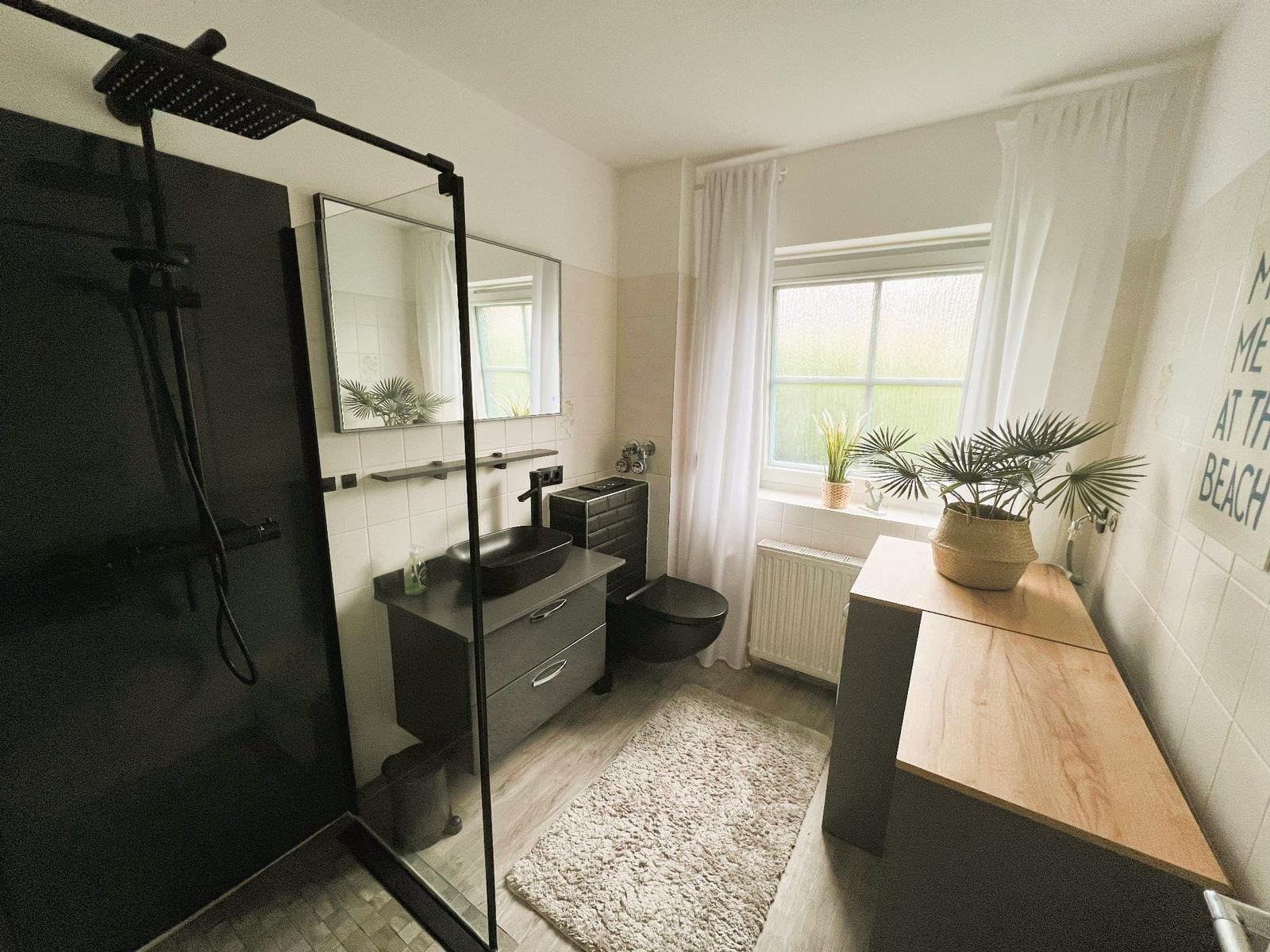 Badezimmer mit Dusche, Waschbecken und Toilette. Fenster mit Pflanzen und Holzoberfläche.