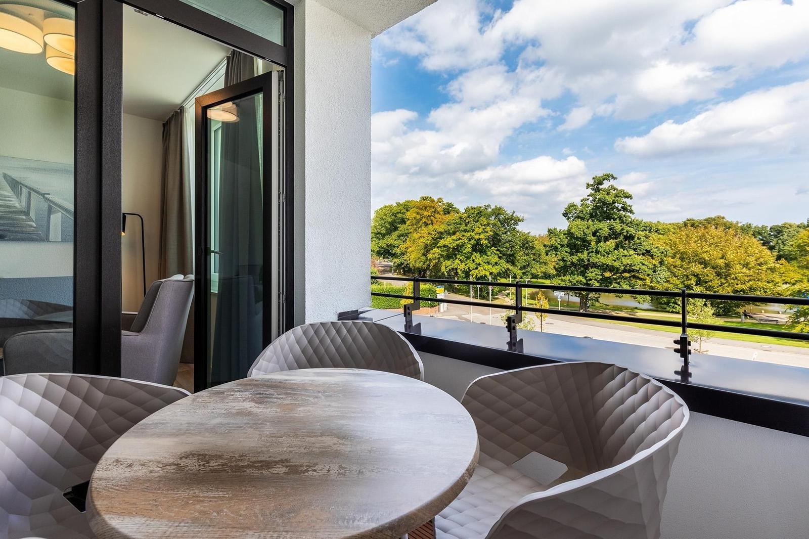 Terrasse mit Tisch und Stühlen. Blick auf grüne Bäume und einen See.