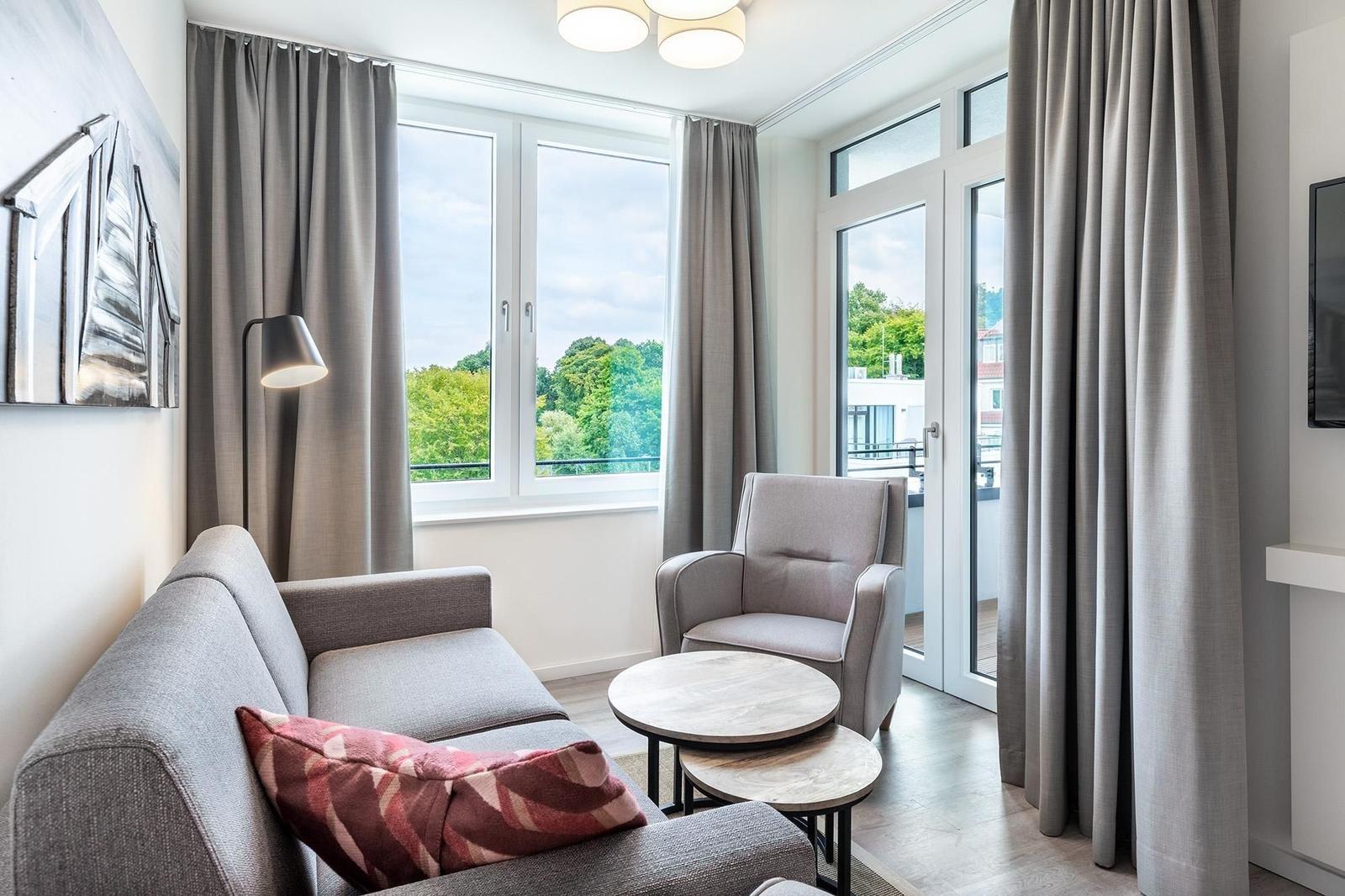 Wohnzimmer mit Sofa, Sessel und Tisch. Fenster und Balkontür bieten Blick auf Grün.