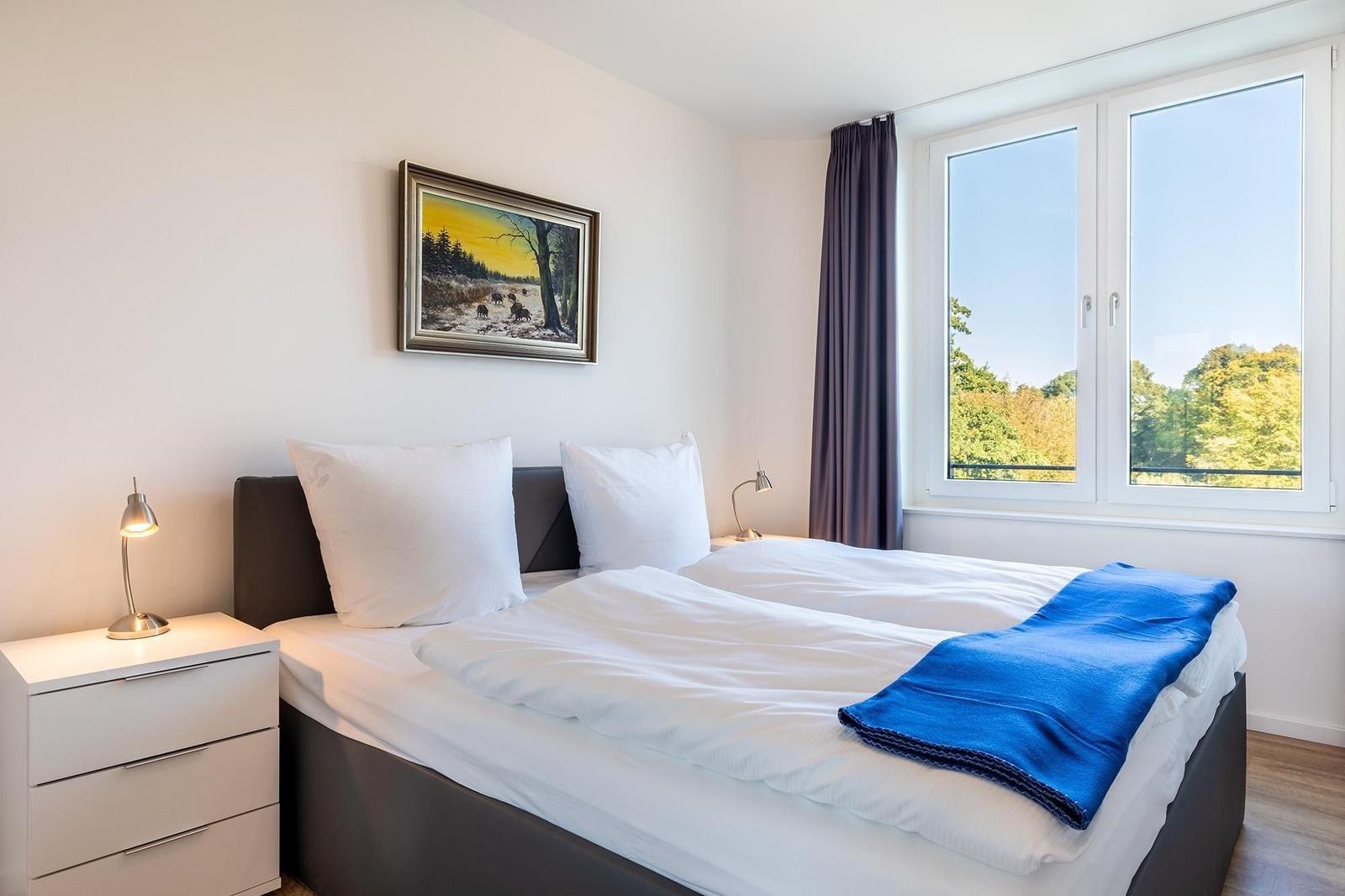 Schlafzimmer mit Bett, Nachttisch und Fensterblick auf Wald.