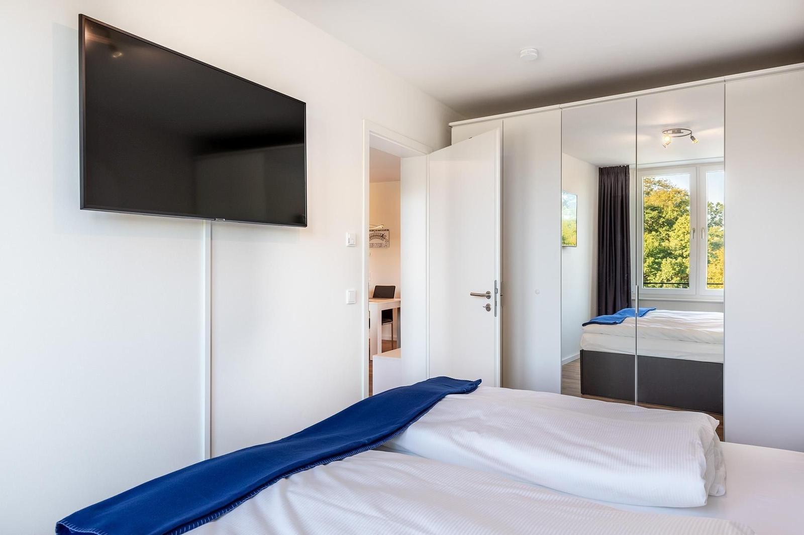 Schlafzimmer mit Bett, TV und Spiegelschrank. Durchgang zu Arbeitsbereich.