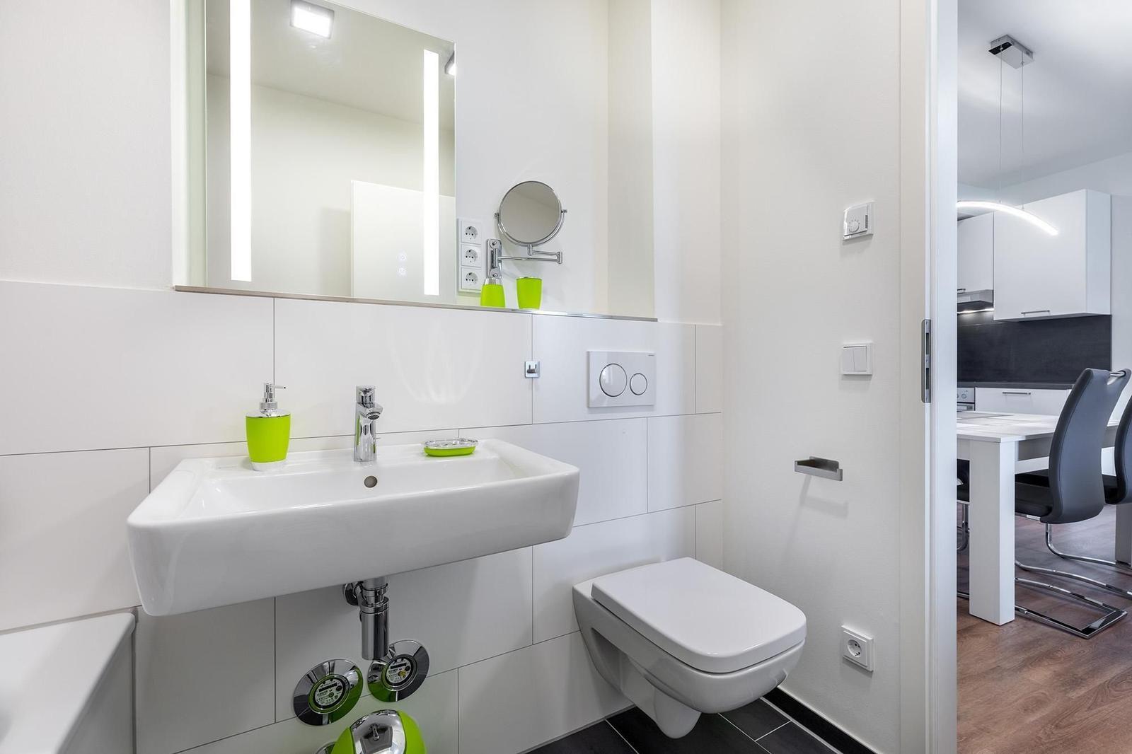 Badezimmer mit Waschbecken, Spiegel und Toilette. Grüne Accessoires sichtbar.