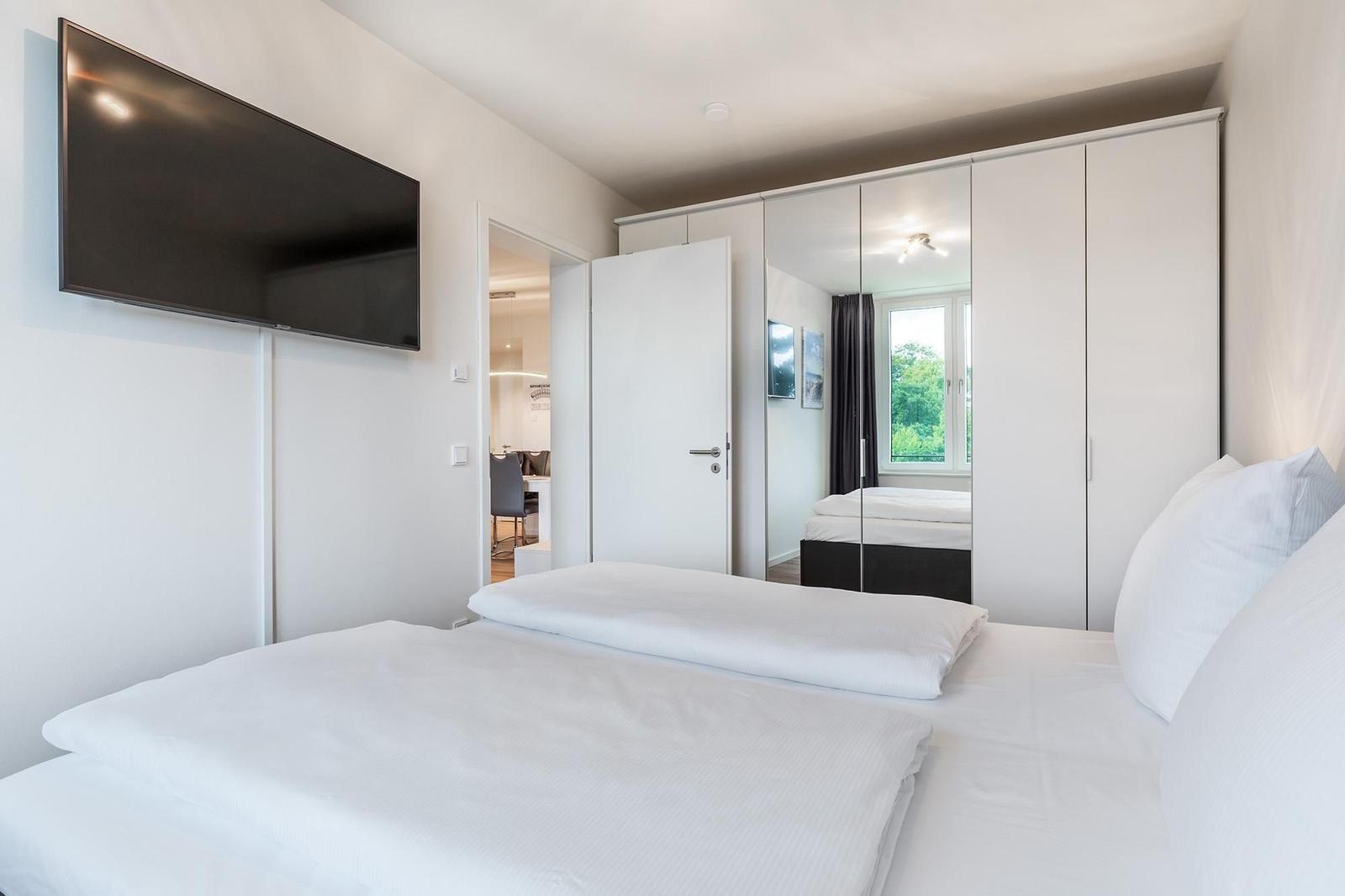 Schlafzimmer mit Bett, Spiegelschrank und Wandfernseher. Blick in die Küche.