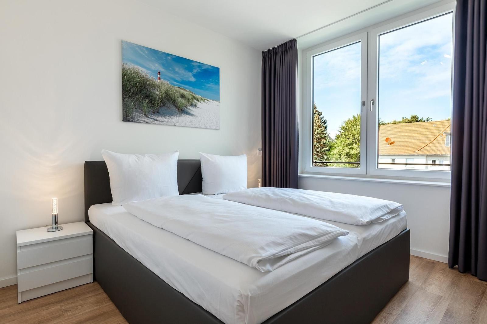 Schlafzimmer mit Doppelbett, Nachttisch und Fensterblick auf Dach und Bäume.