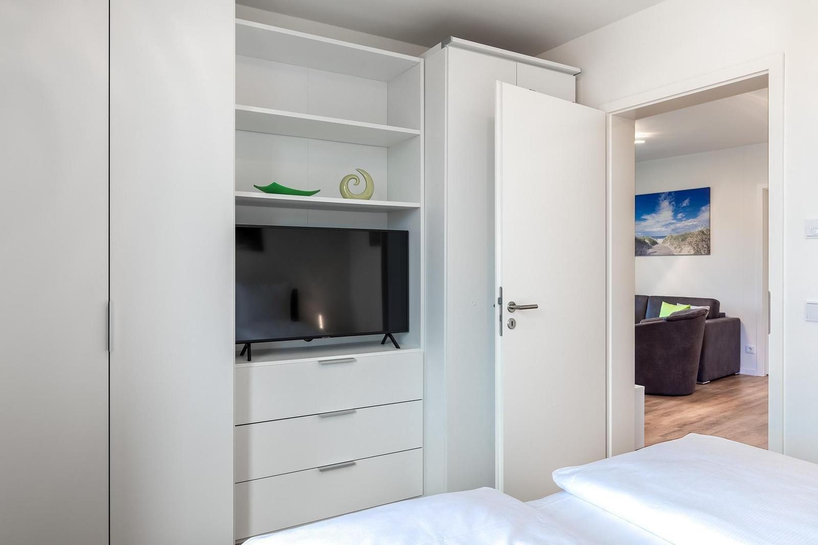 Schlafzimmer mit weißem Schrank, Fernseher und Blick in Wohnzimmer.