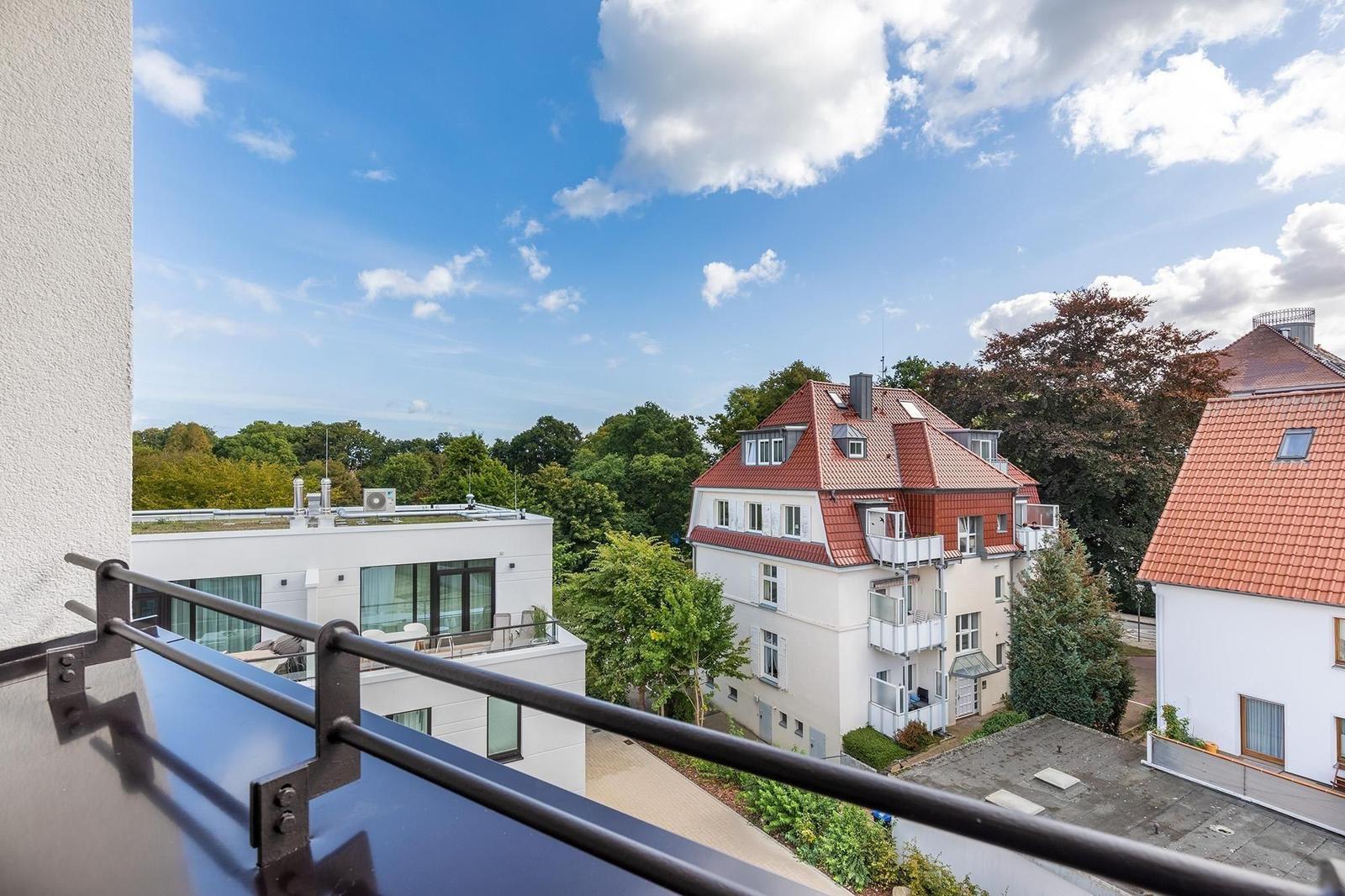 Blick vom Balkon auf Wohnhäuser und Bäume unter blauem Himmel mit weißen Wolken.