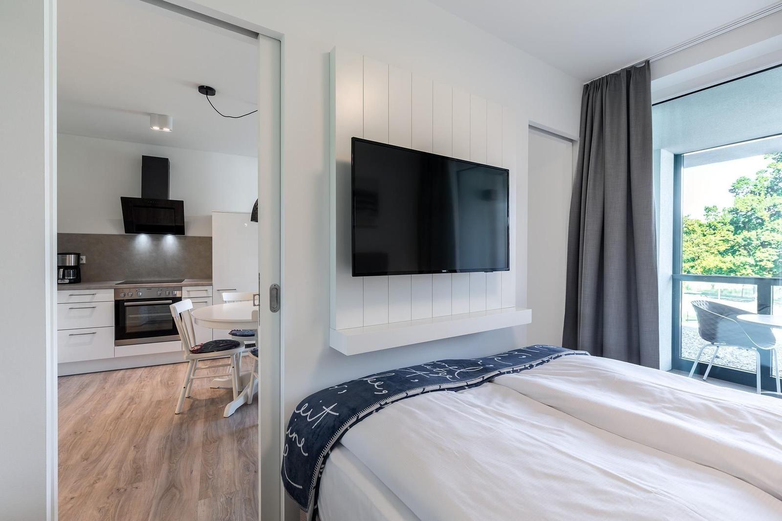 Schlafzimmer mit TV und Blick auf Balkon. Küche im Hintergrund sichtbar.