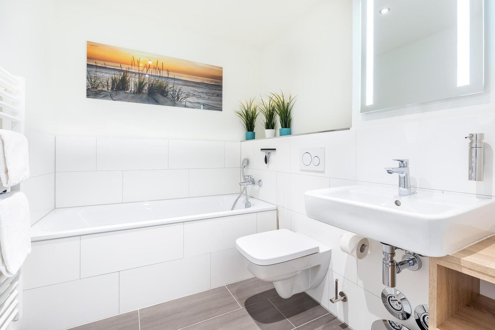 Badezimmer mit Badewanne, Waschbecken und Toilette. Wandbild und Pflanzen sichtbar.