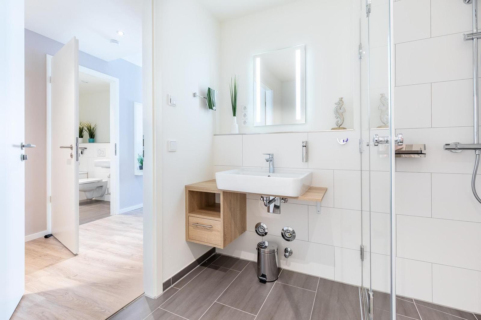 Badezimmer mit Dusche, Waschbecken und Spiegel. Tür führt in Toilettenraum.