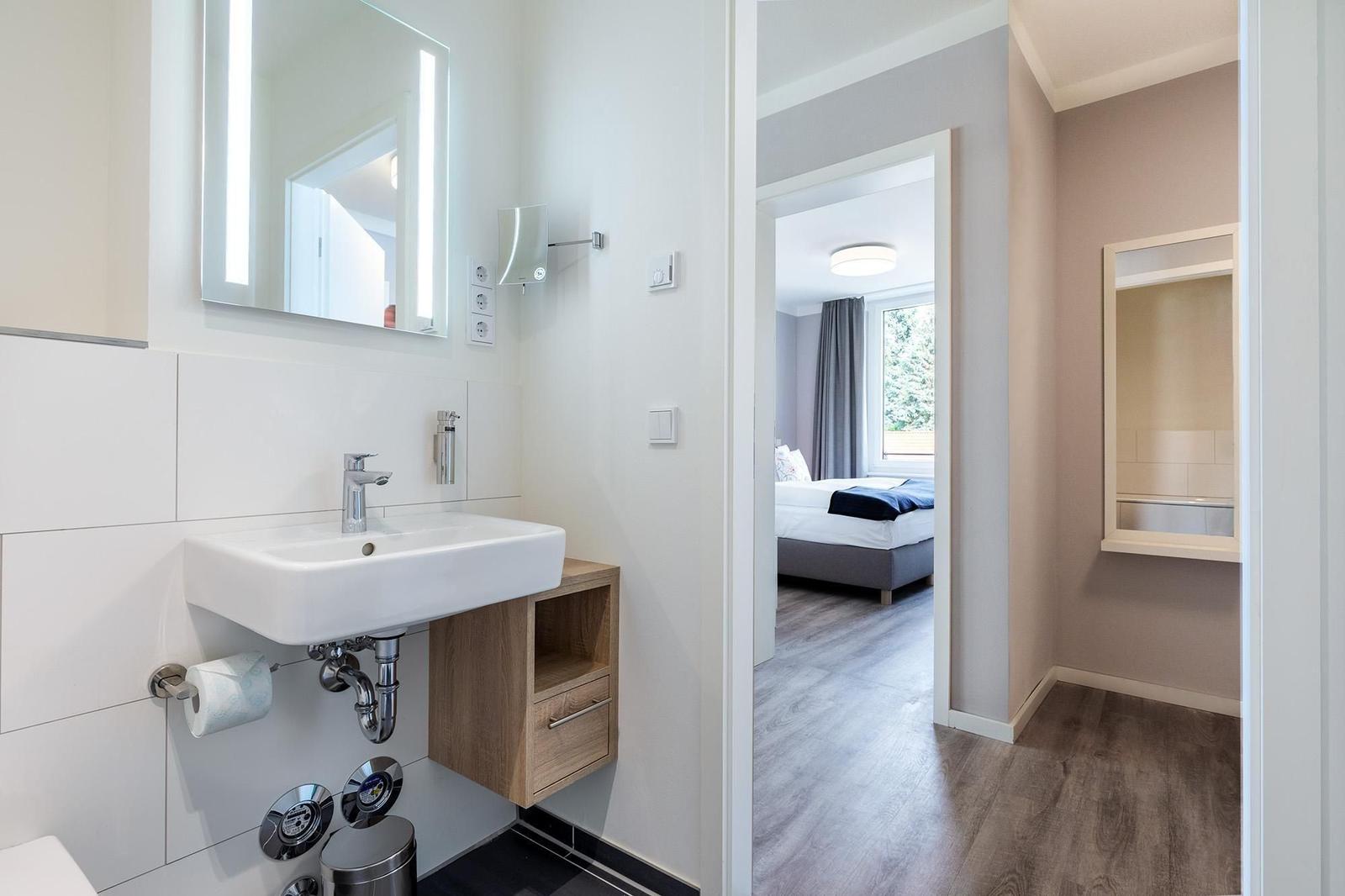 Badezimmer mit Waschbecken und Spiegel. Blick in Schlafzimmer mit Bett und Fenster.